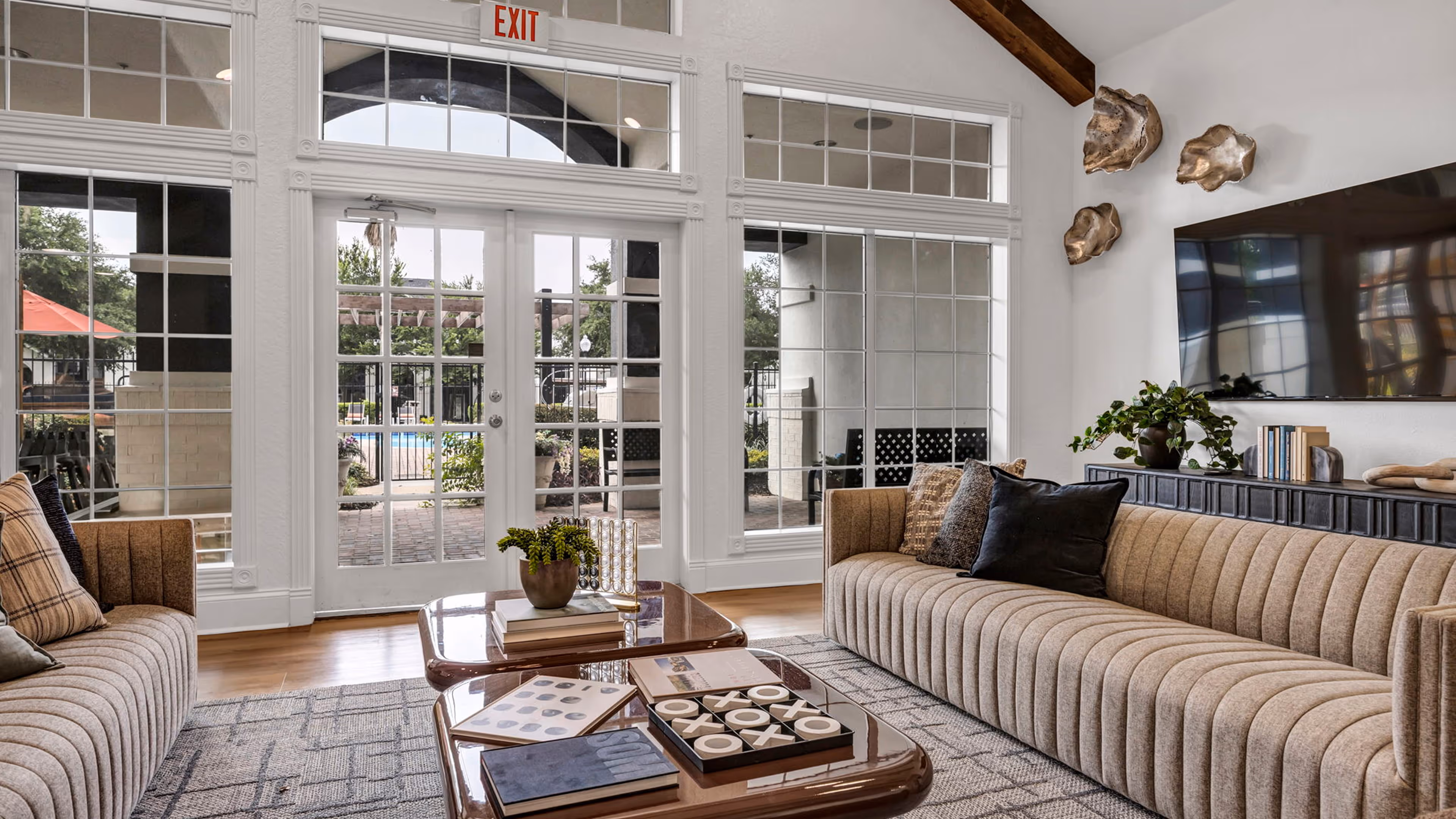 Modern living room with two beige sofas, wooden coffee tables with books and decorations, large windows, and a TV mounted on the wall.